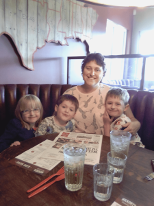 Helen Jones, our Warrior Wednesday spotlight, sits at a restaurant booth with her three children (two boys and a girl). They are all smiling. There's two menus and four glasses of water on the table in front of them.